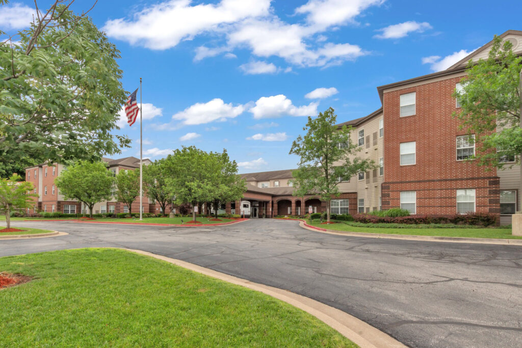 Town Village Tulsa driveway to the dark brick building with trees surrounding it and an American flag, a senior living community near Broken Arrow offering independent living.