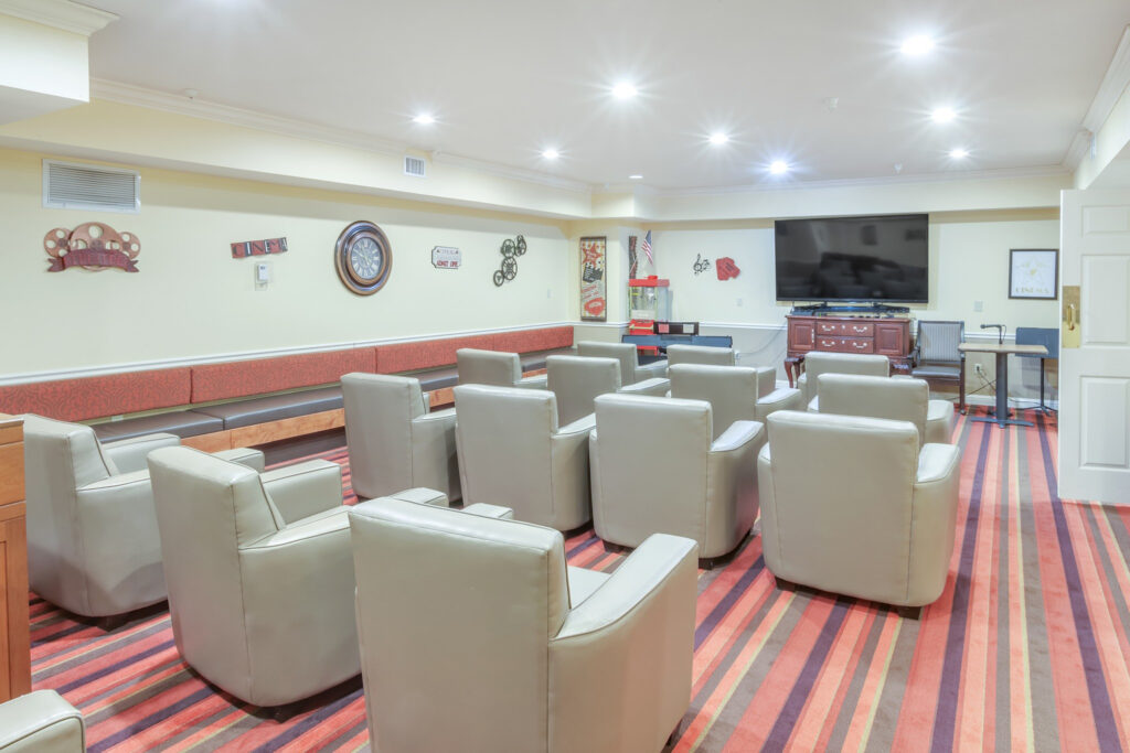 A movie theater room with a striped carpet and rows of gray sofa chairs facing the TV at Town Village Tulsa, a senior living community near Glenpool.