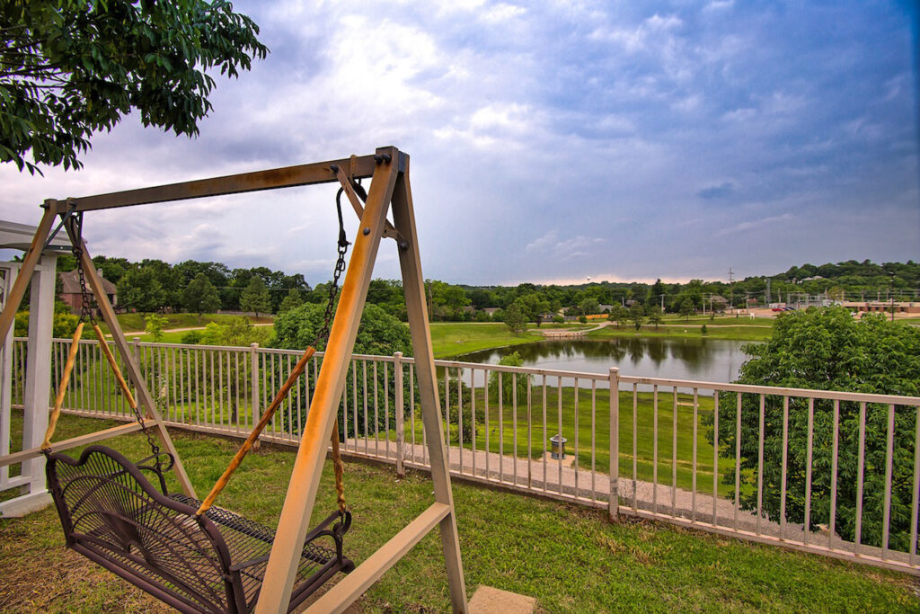 A metal outdoor swing placed in front of a fence, overlooking a beautiful view of the green scenery and lake at Town Village Tulsa, a senior living community near Glenpool.