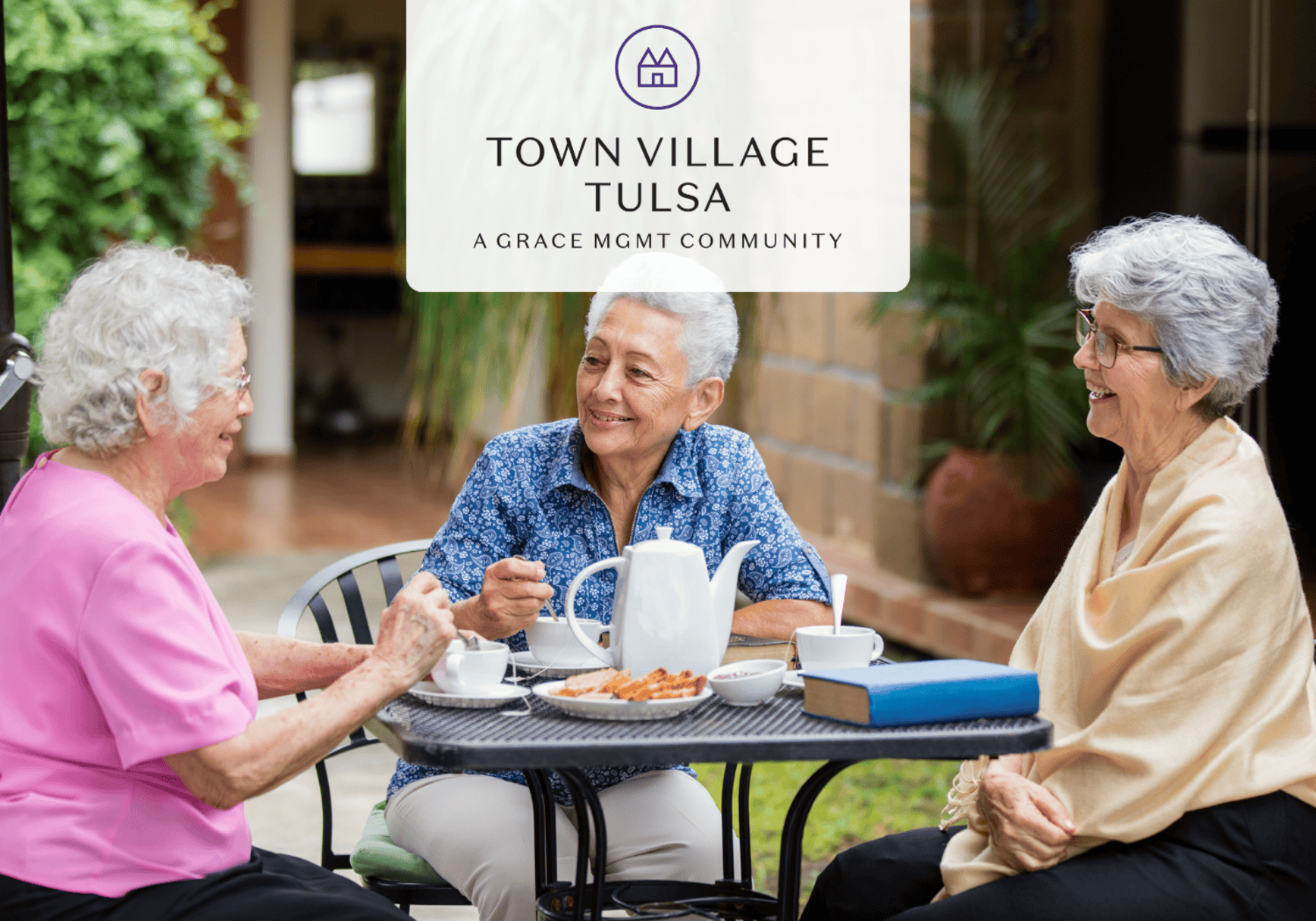 Three elderly women sit at a round outdoor table enjoying tea and snacks, smiling and chatting, with a Town Village Tulsa sign visible above them (A Grace Mgmt Community).