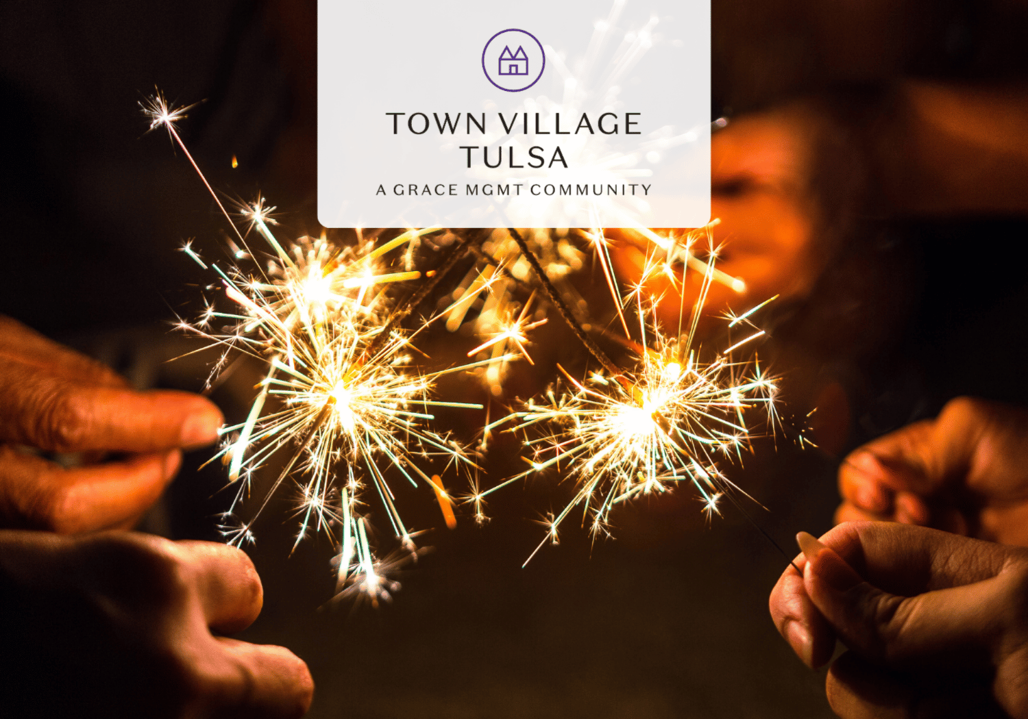 Hands holding lit sparklers in a dark scene, bright sparks fly with a pale logo overlay reading Town Village Tulsa at the center