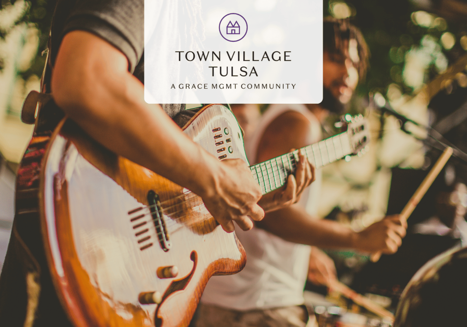 Close-up of a guitarist playing an orange electric guitar at a live outdoor performance, with a second guitarist in the background and a translucent Town Village Tulsa logo overlay.