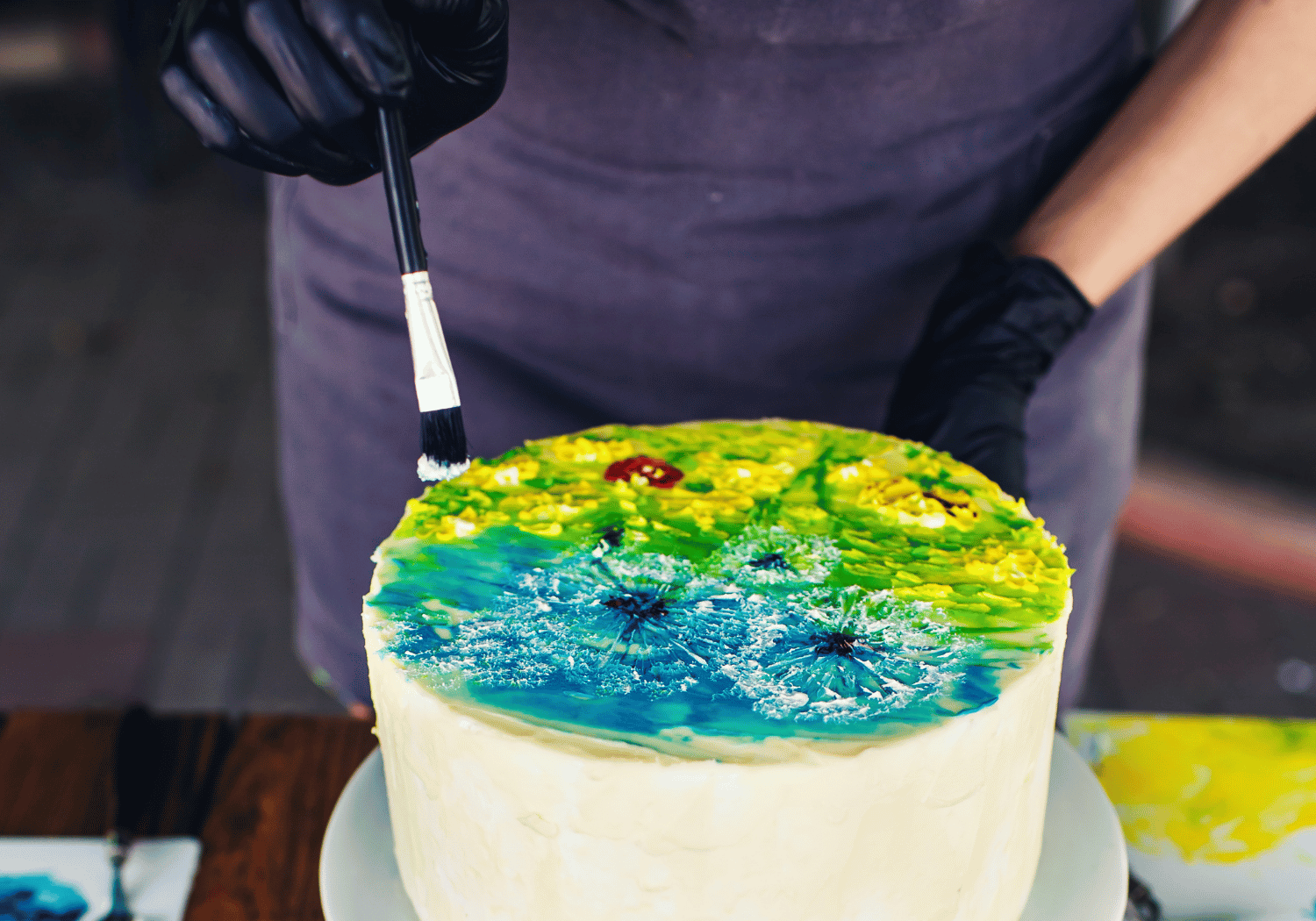 Person wearing black gloves drags a brush over a rainbow-colored cake, adding blue, green, and yellow icing.
