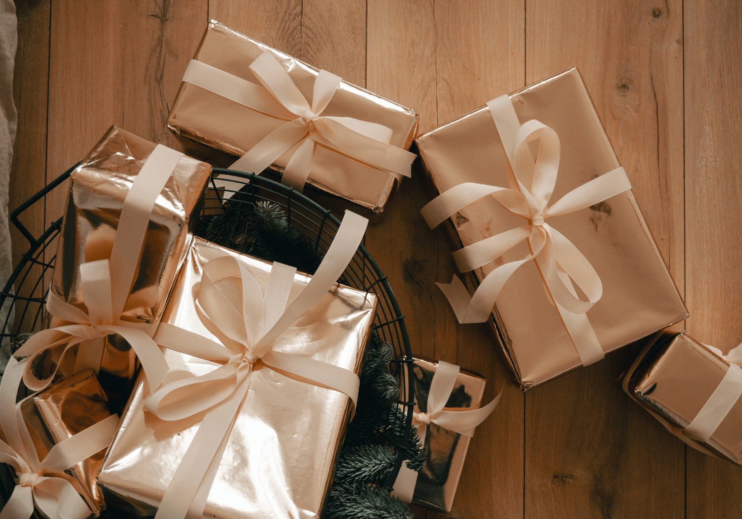Gold-wrapped gift boxes tied with white ribbons arranged on a wooden floor, with a basket and evergreen sprigs nearby.