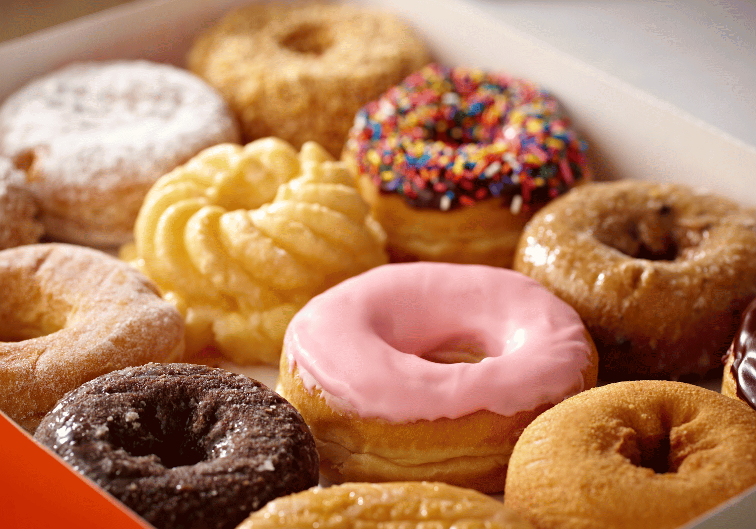 Assorted donuts in a box, including pink frosted ring, chocolate glaze, sprinkled, and powdered sugar varieties